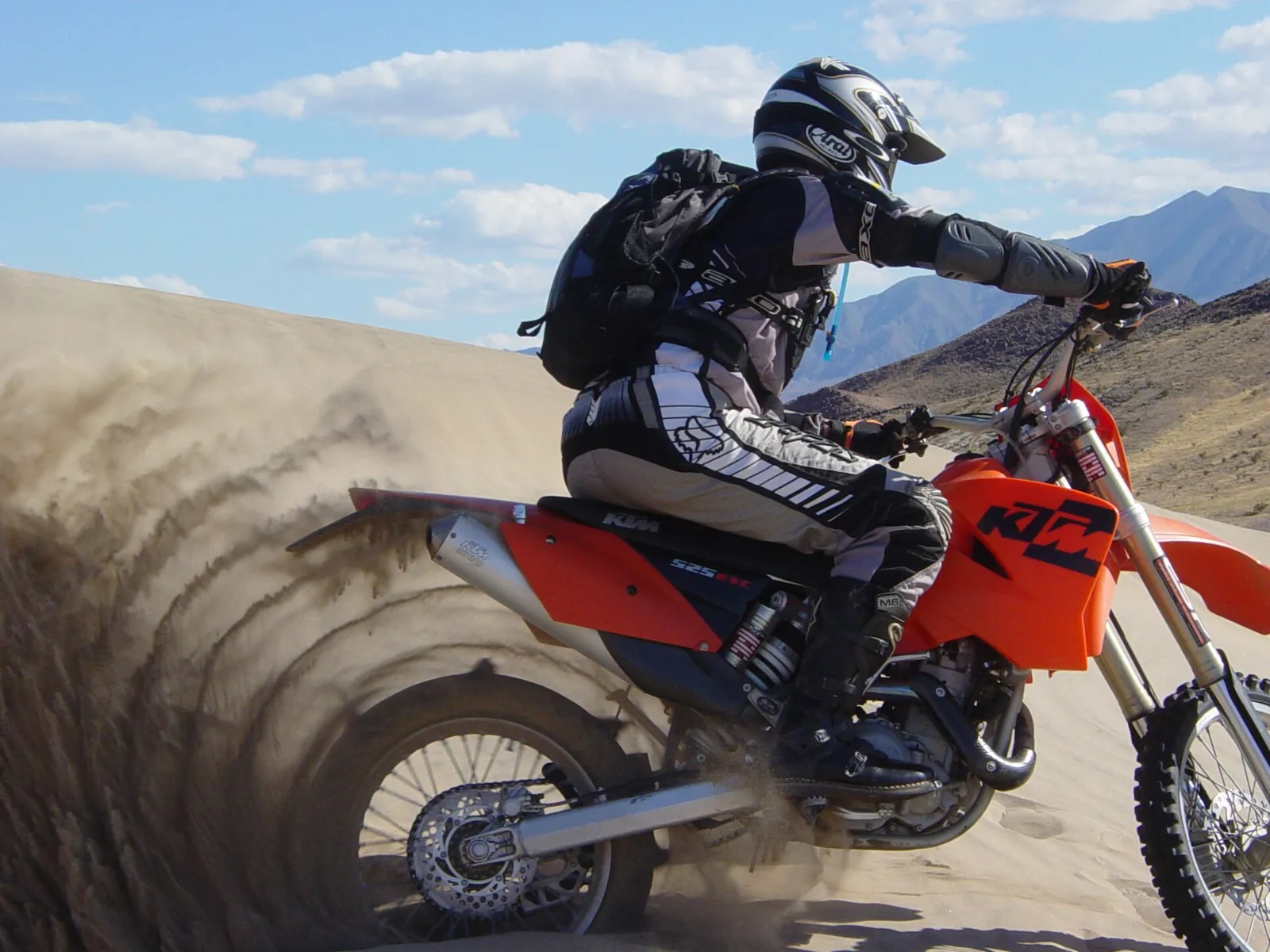 Pilota di motocross in sella a una KTM che affronta una duna di sabbia sollevando sabbia